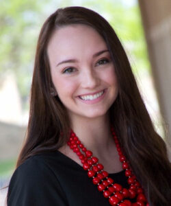 Young woman with long brown hair weraing a large red beaded necklace and black blouse. Photographed outside